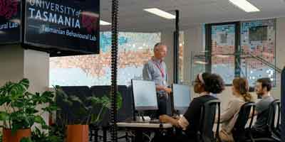 A group of individuals working at computers in an office with large windows displaying the University of Tasmania signage.
